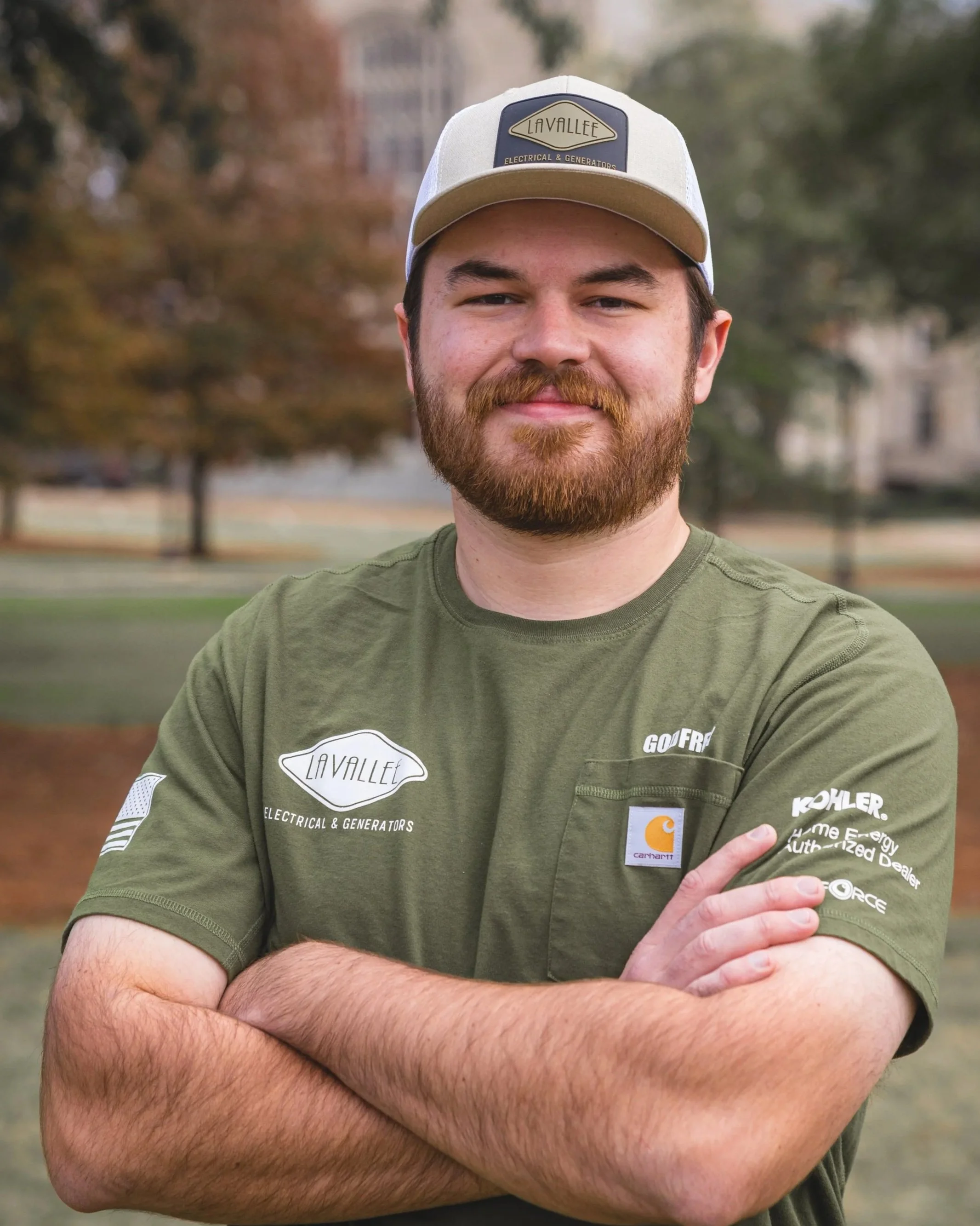 A man with a beard and mustache is standing outdoors with arms crossed, wearing a green t-shirt with logos and a beige and gray baseball cap, in a park with trees and buildings in the background.