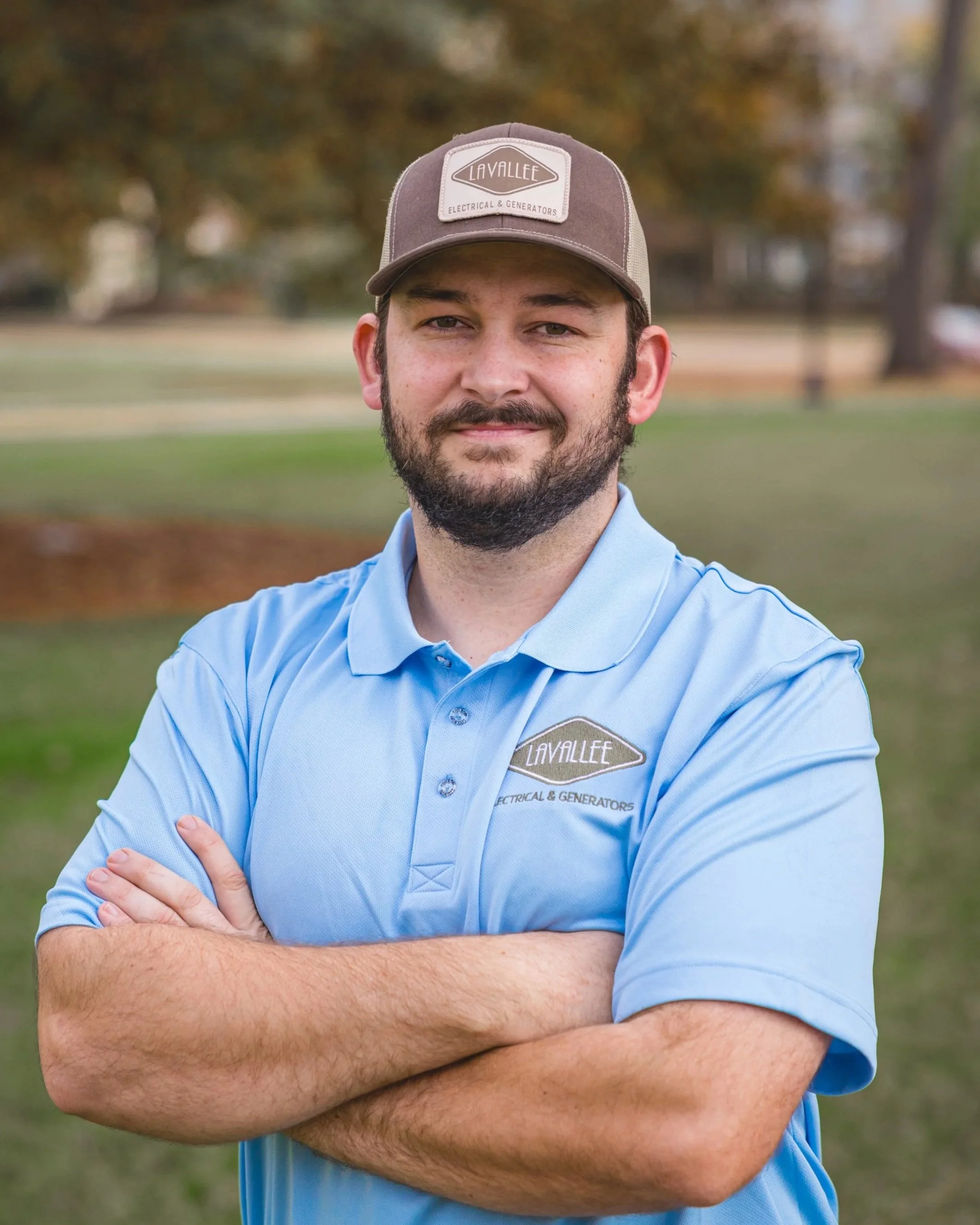 A man with a beard in a light blue polo shirt and a brown baseball cap, standing outdoors with his arms crossed, in a park during autumn.