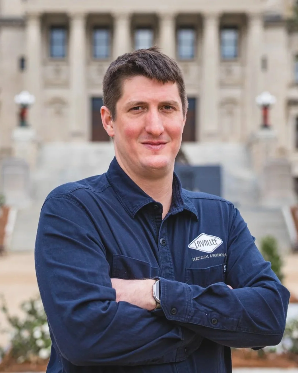 A man with short brown hair and a slight smile crossing his arms, wearing a navy blue work shirt with a patch that reads "LAVALLEE Electrical & Generators," standing outdoors in front of a historic stone building with stairs and lampposts.