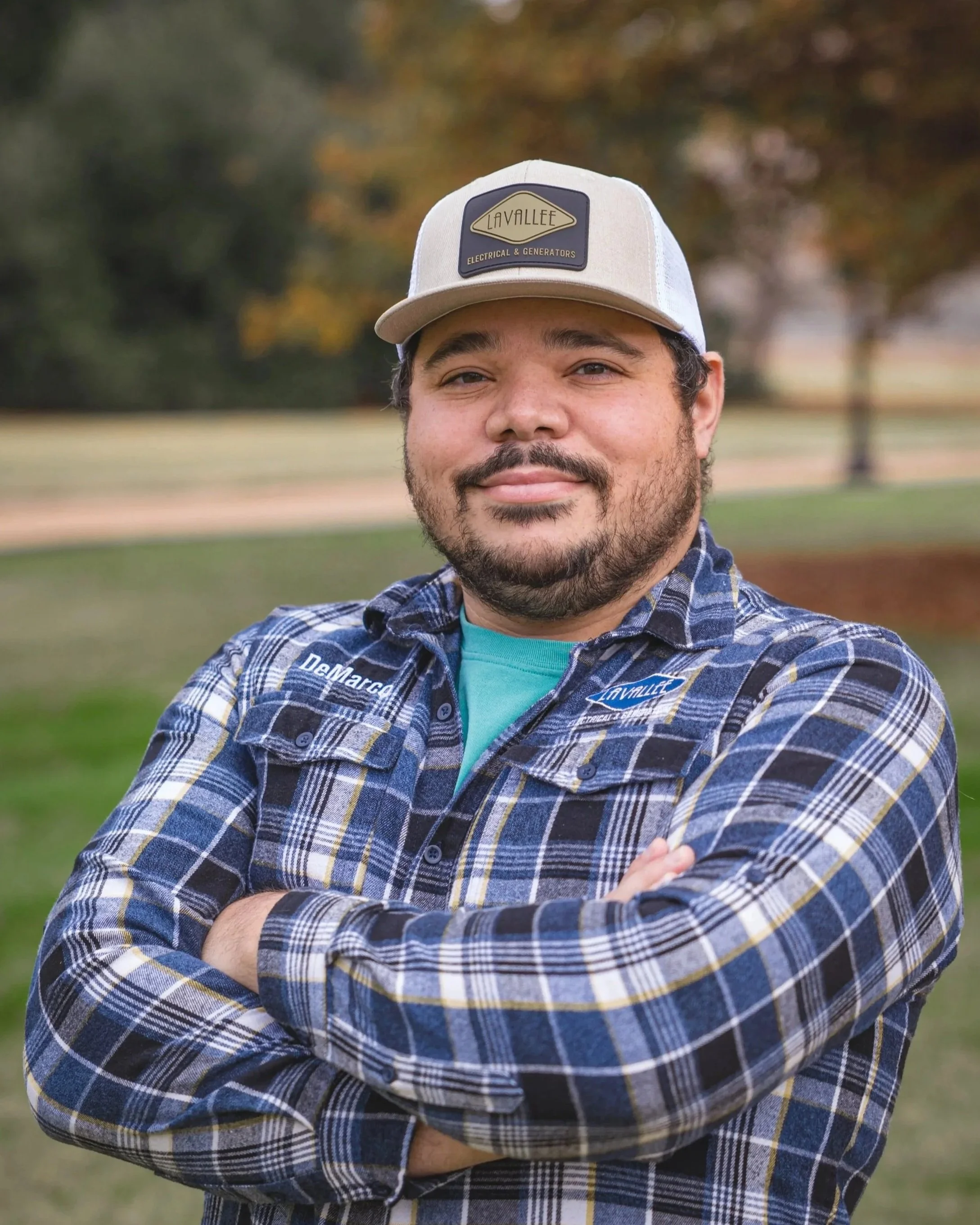 A man with a beard and mustache wearing a plaid shirt and a cap with a logo, standing outdoors with trees and grass in the background.