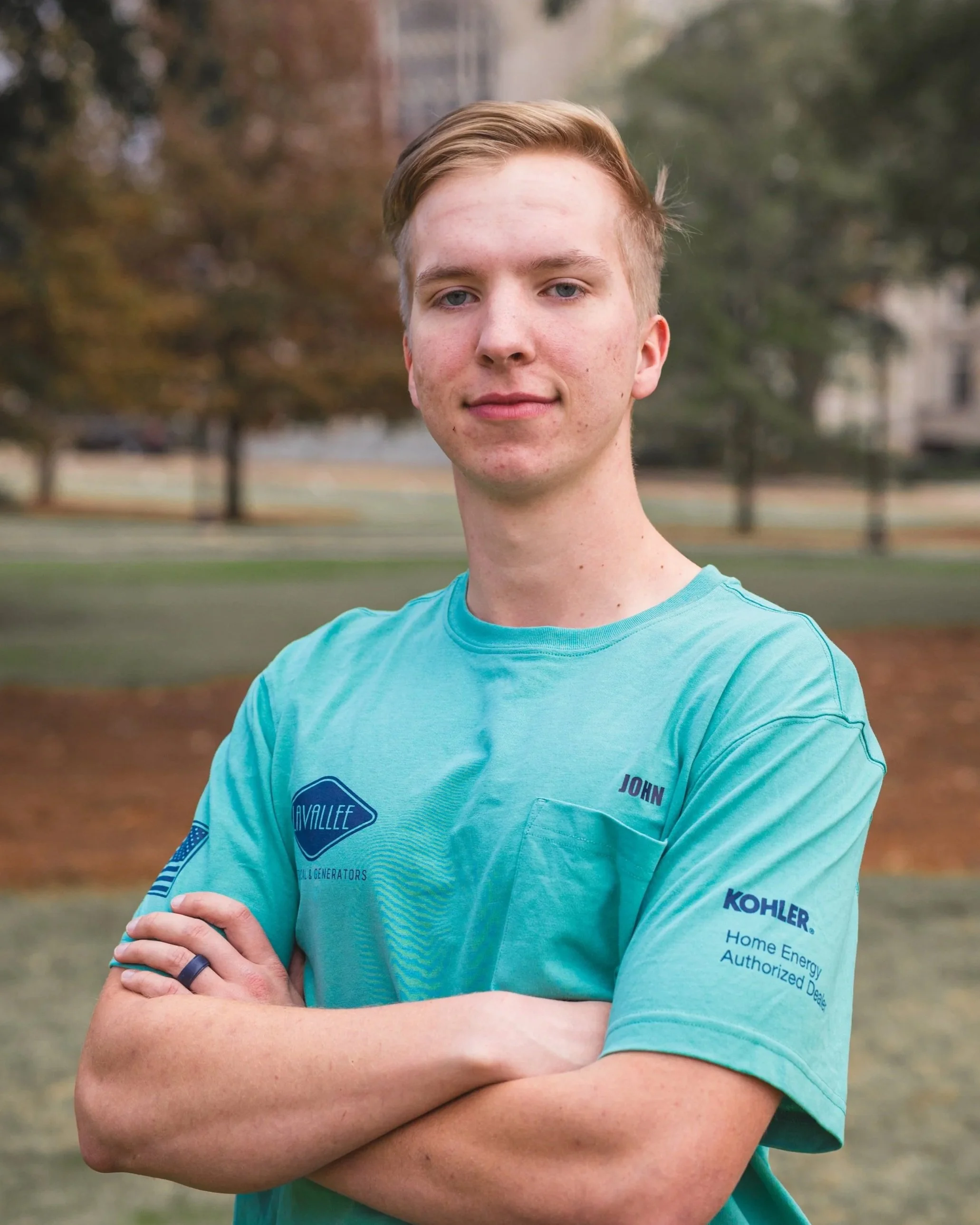 A young man with blond hair and blue eyes standing outdoors with arms crossed, wearing a teal T-shirt with sponsor logos and the name 'John' on it, in a park-like setting with blurred trees and grass in the background.