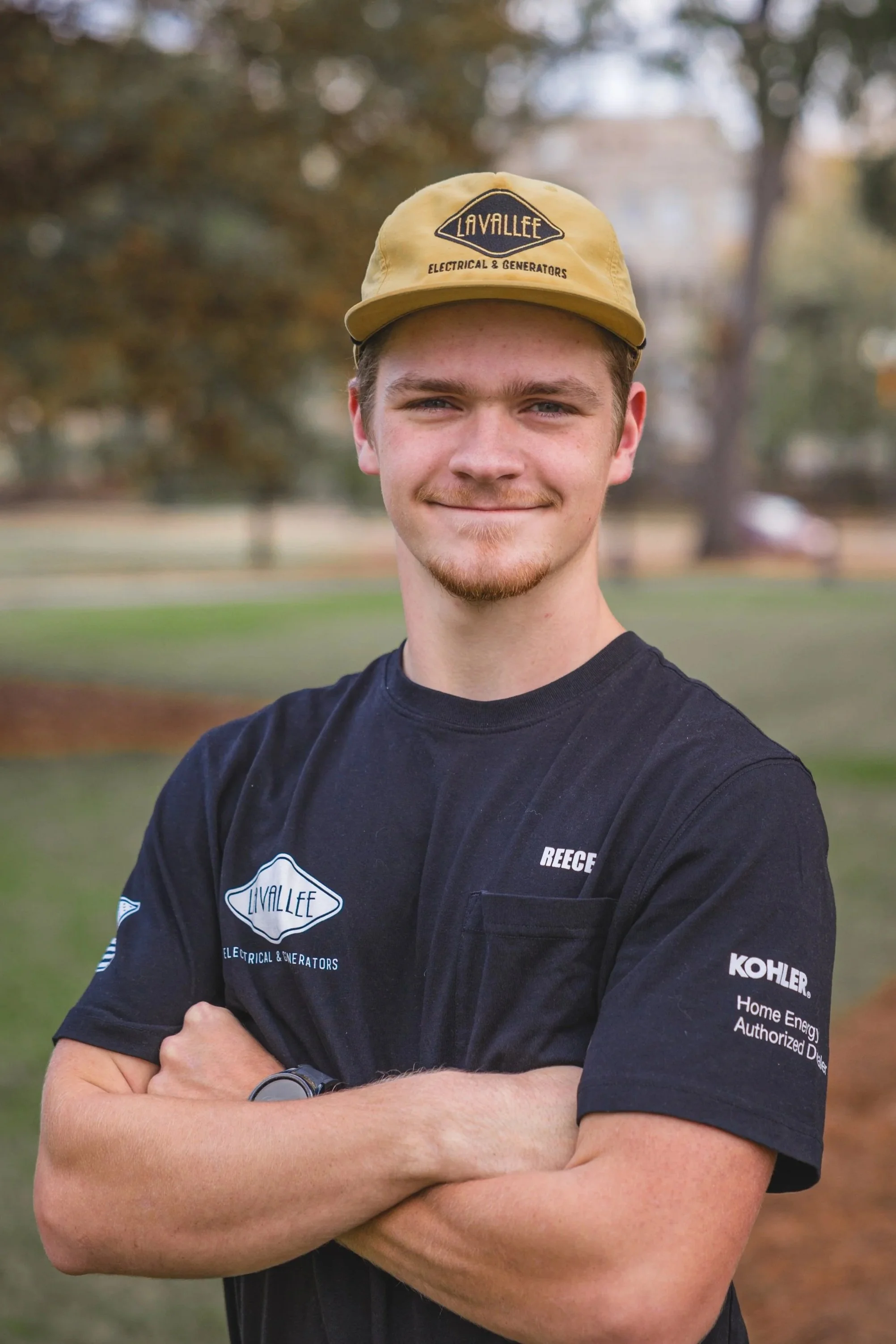 Young man in black work shirt and yellow cap standing outdoors with arms crossed