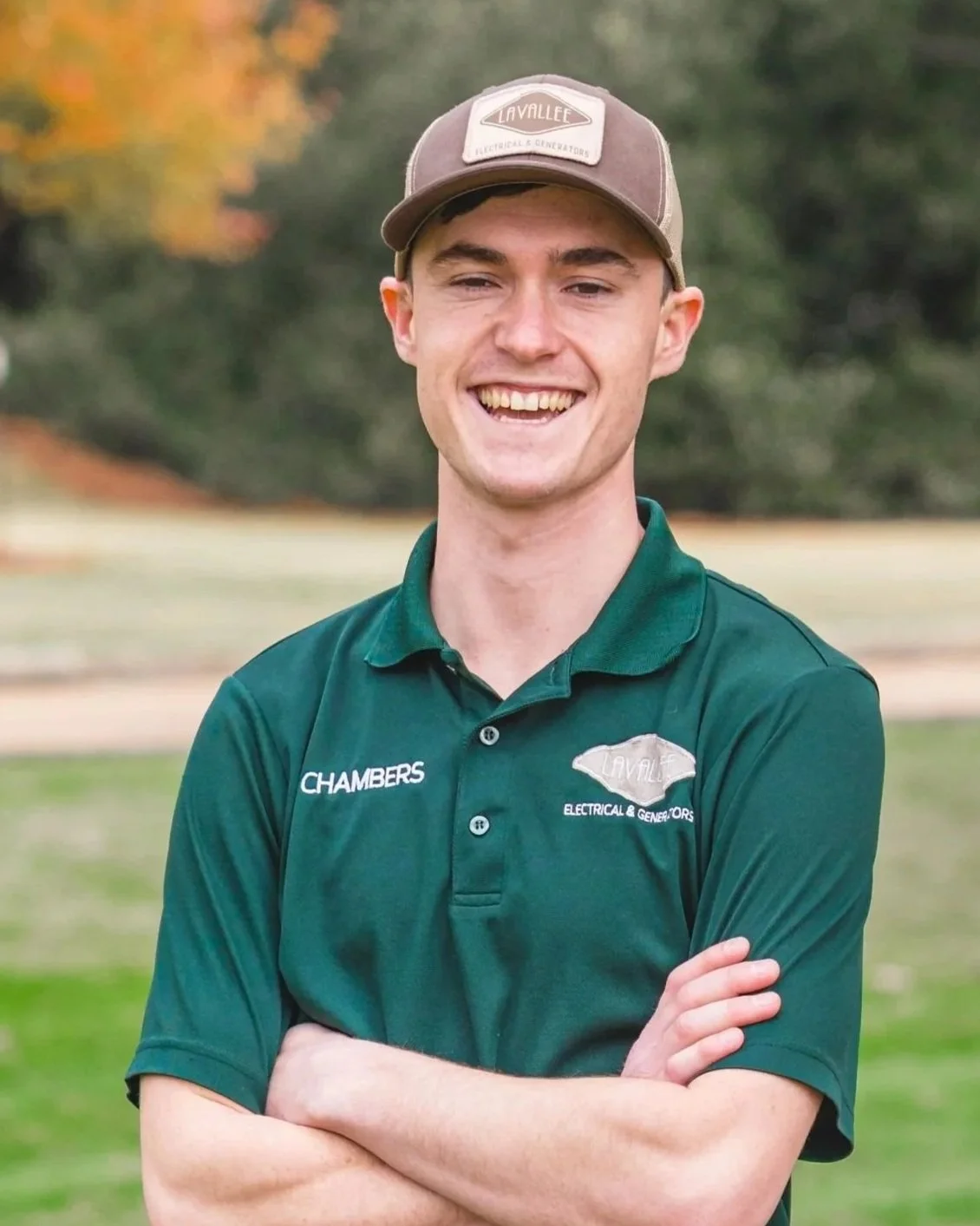 A young man smiling outdoors, wearing a green polo shirt with company logos for 'Lavallee Electrical & Generators' and 'Chambers', and a baseball cap with the same company branding, standing with arms crossed in a park-like setting with blurred trees in the background.