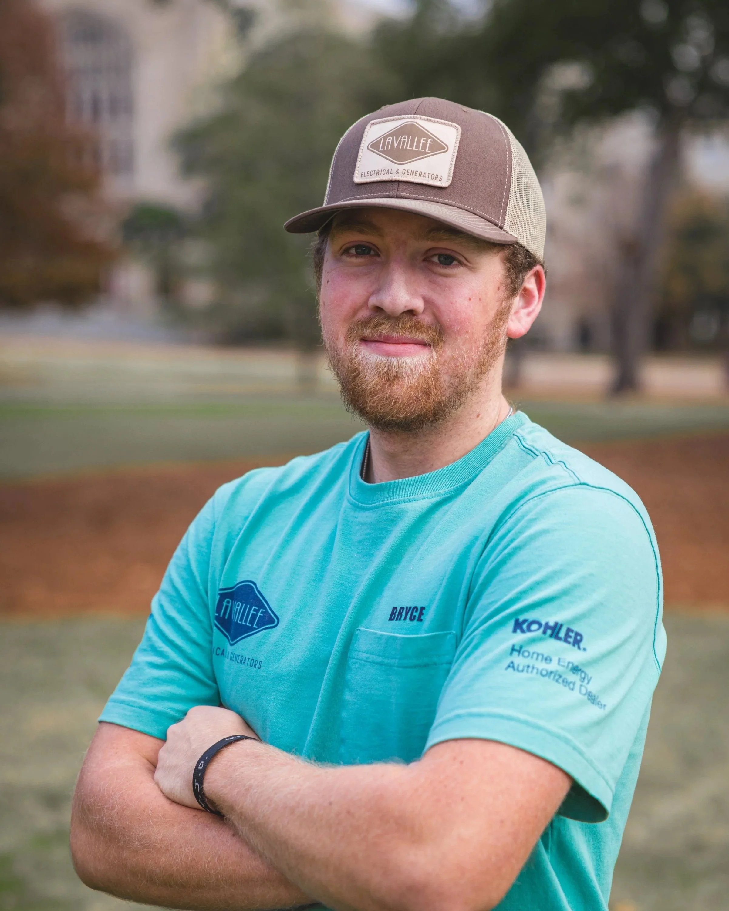 A young man with a beard and mustache wearing a baseball cap, a teal T-shirt with logos and text, and a black wristband, standing outdoors in a park with blurred trees and grass in the background.