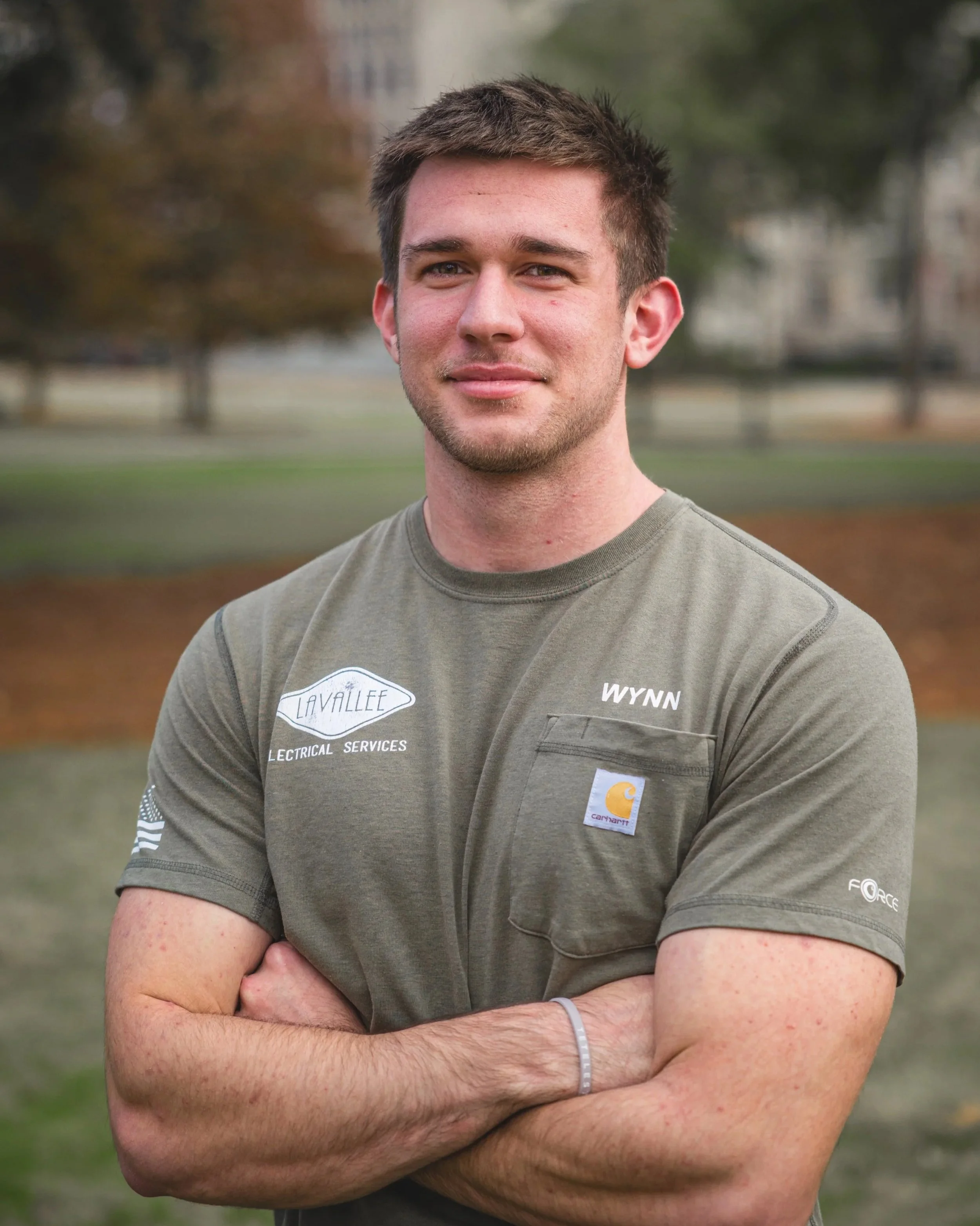 A young man with short brown hair and light skin, standing outdoors with arms crossed, smiling at the camera. He's wearing a gray T-shirt with various logos and text, including a Carhartt logo and the name 'Wynn,' in a park-like setting with trees and grass in the background.