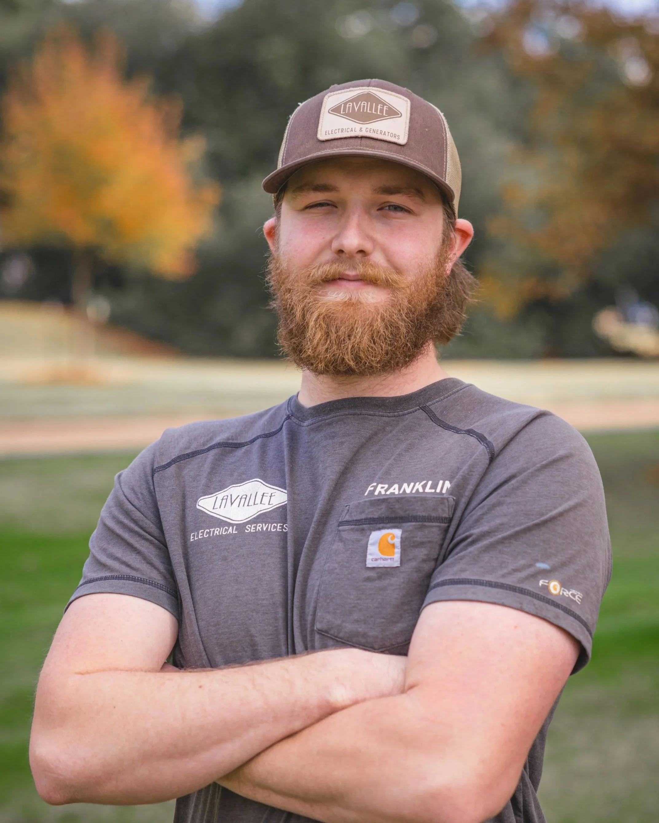 A young man with a red beard and wearing a brown and tan baseball cap and a gray work shirt with patches, standing outdoors with arms crossed, trees with autumn leaves in the background.