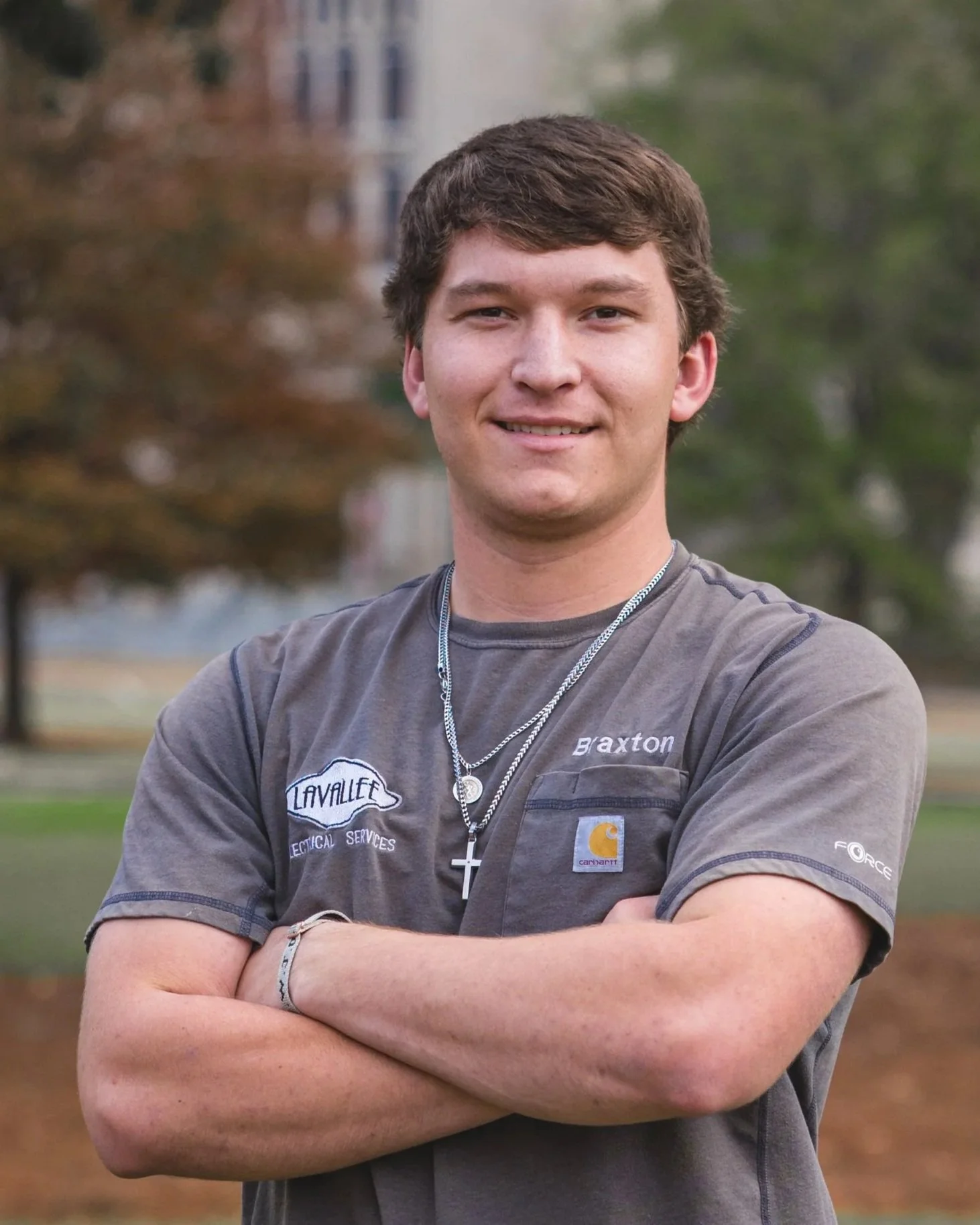 A young man with short brown hair standing outdoors with arms crossed, smiling at the camera, with trees and a building in the background.