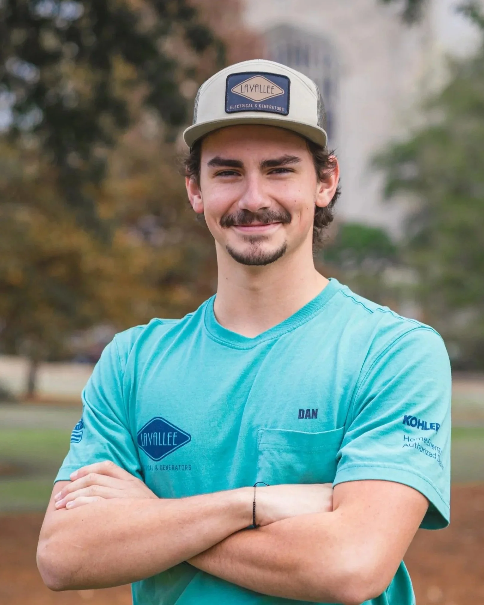 A young man with dark hair and a beard, smiling with arms crossed, wearing a light teal shirt and a beige cap with a decal that says 'LAVALLEE Electrical & Generators', standing outdoors in a park.