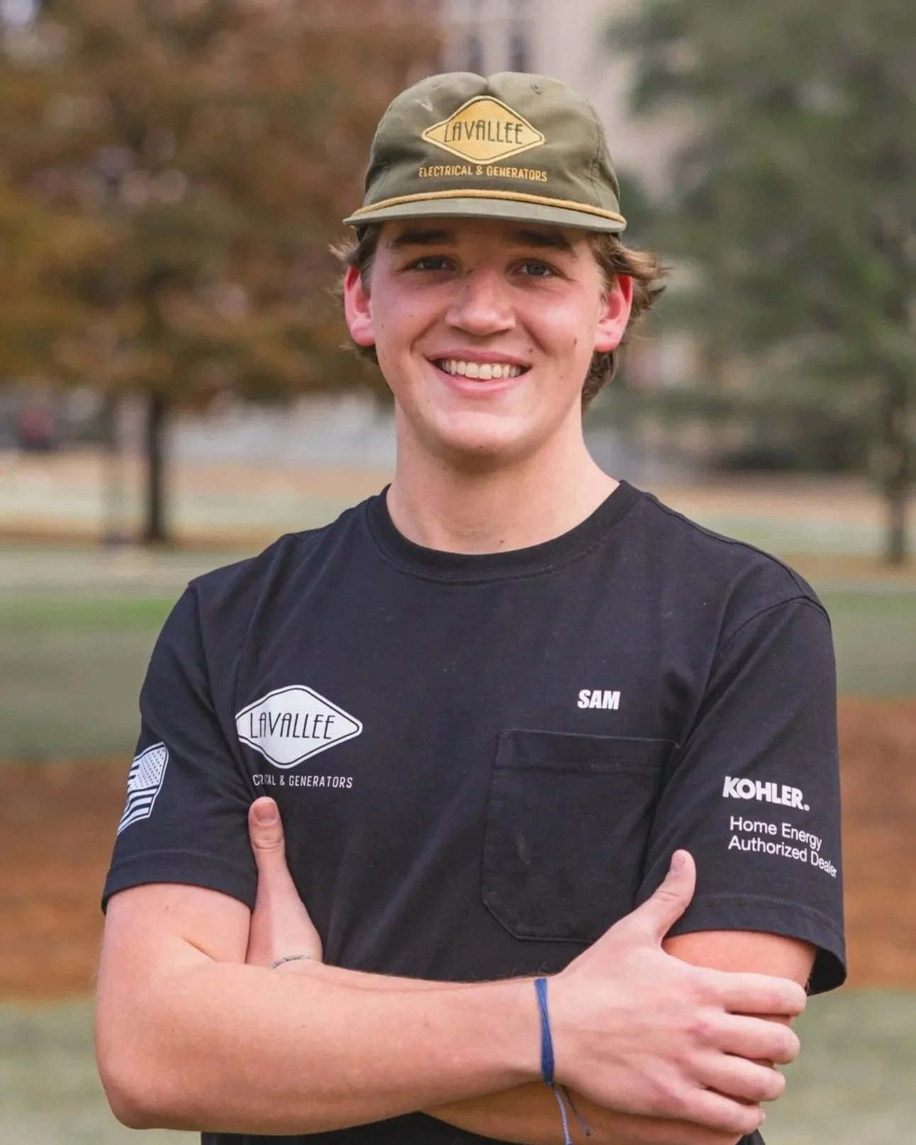 Young man smiling outdoors, wearing a black t-shirt with logos and a tan baseball cap, crossing his arms with hands on his upper arms, background of trees and grass.