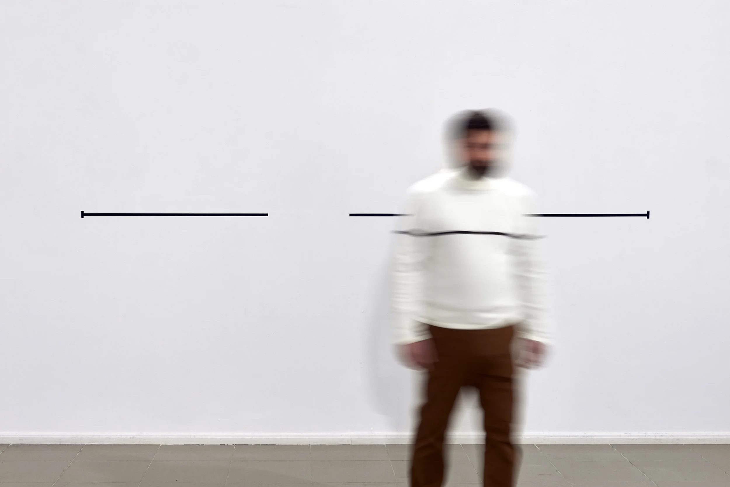 A blurry man standing in front of a plain white wall with black lines.