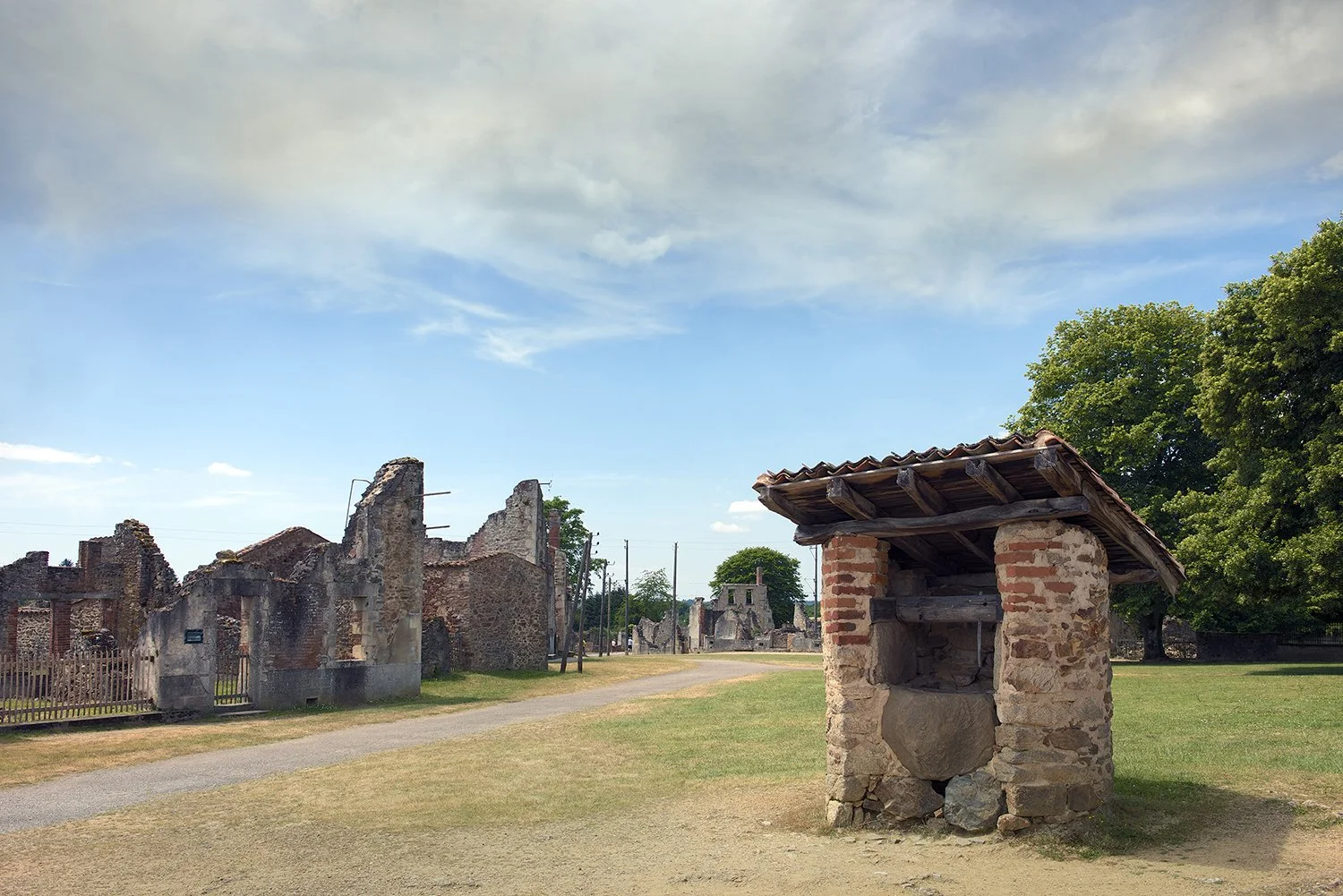 Ruins of an old stone and brick building with a small stone structure in the foreground, green grass, large green trees, and a partly cloudy blue sky.