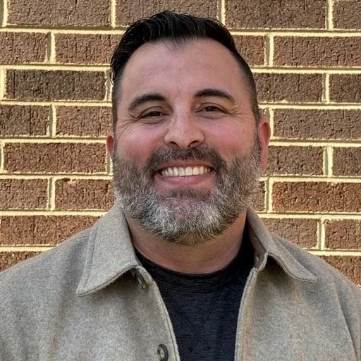 Man with dark hair and beard smiling, wearing a gray jacket in front of a brick wall.