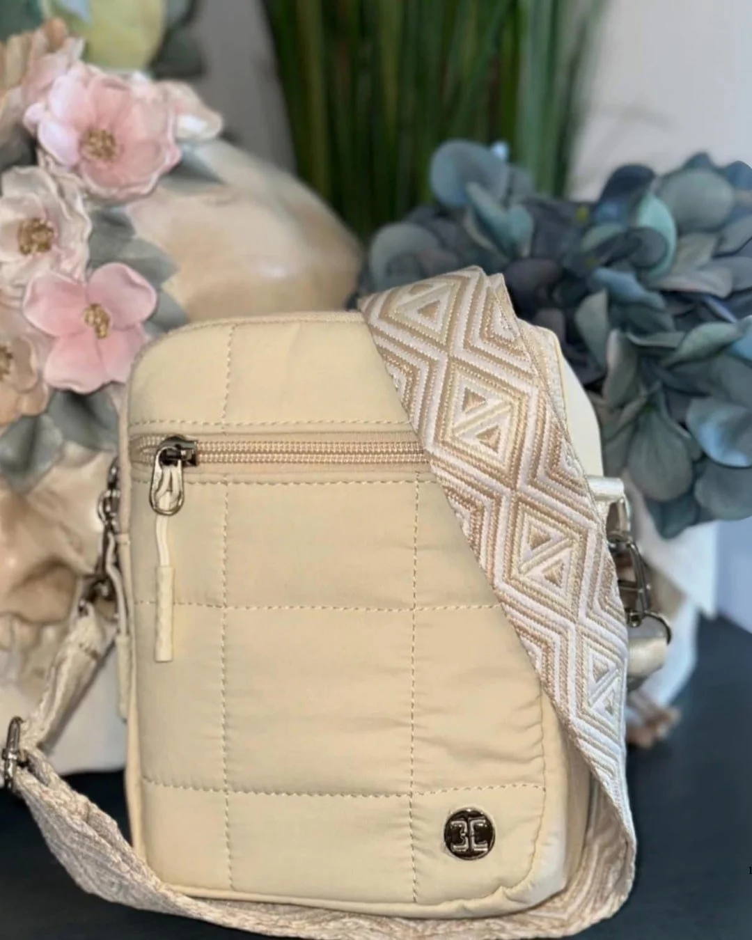 Cream-colored quilted crossbody bag with a geometric patterned strap, silver hardware, and a blurred floral background.