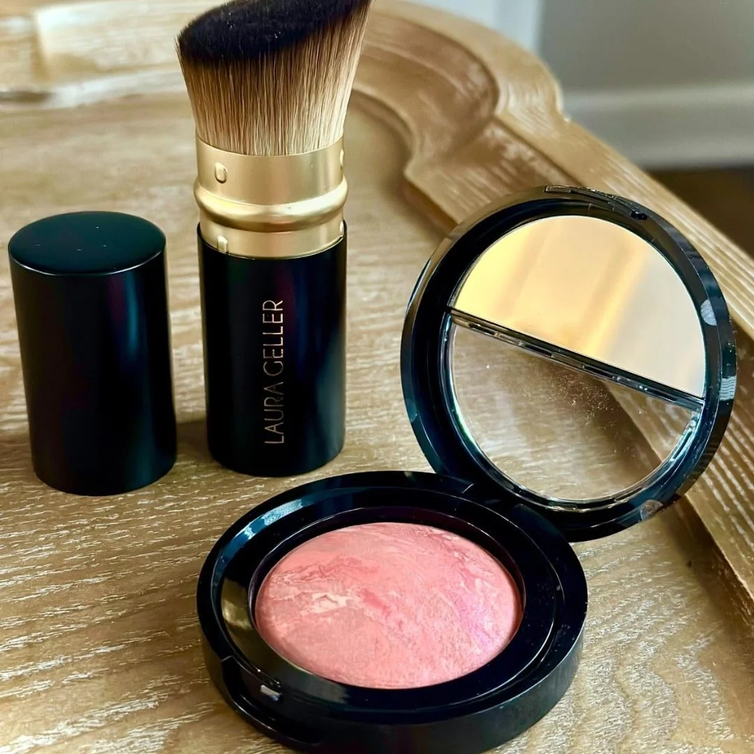Laura Geller retractable makeup brush with black and gold design, paired with two open compacts featuring marbled pink blush and a mirror, displayed on a light wooden surface.