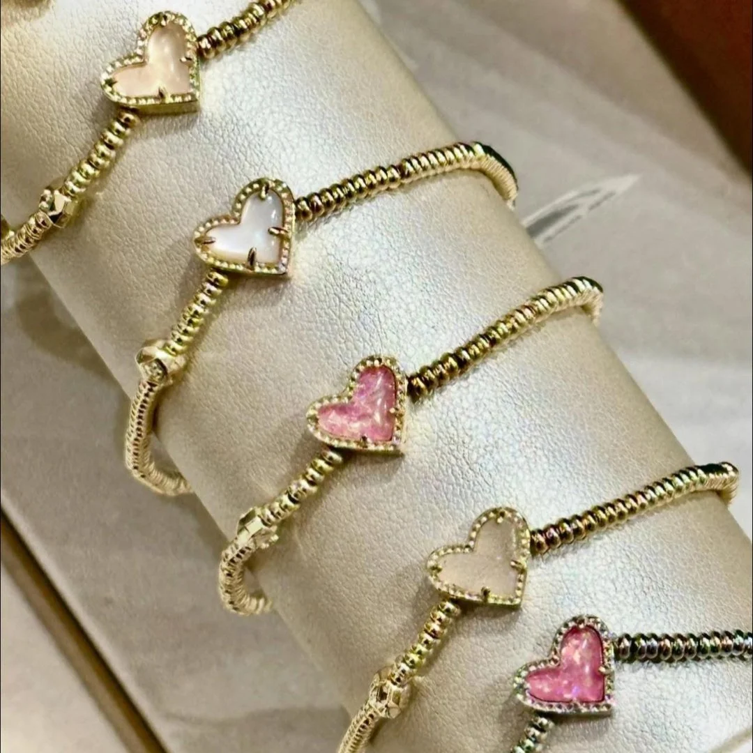 Kendra Scott gold bracelets with heart-shaped charms featuring pearlescent and pink inlays, elegantly displayed on a light cylindrical stand in a retail setting.