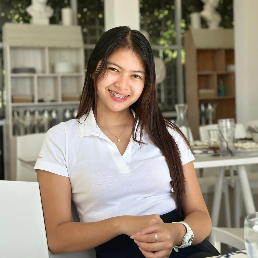 Kristine, website manager for Nicole Fabulous Finds, smiling brightly with braces, seated at a table in a bright cafe, wearing a white polo shirt, with shelves, windows, and greenery in the background.