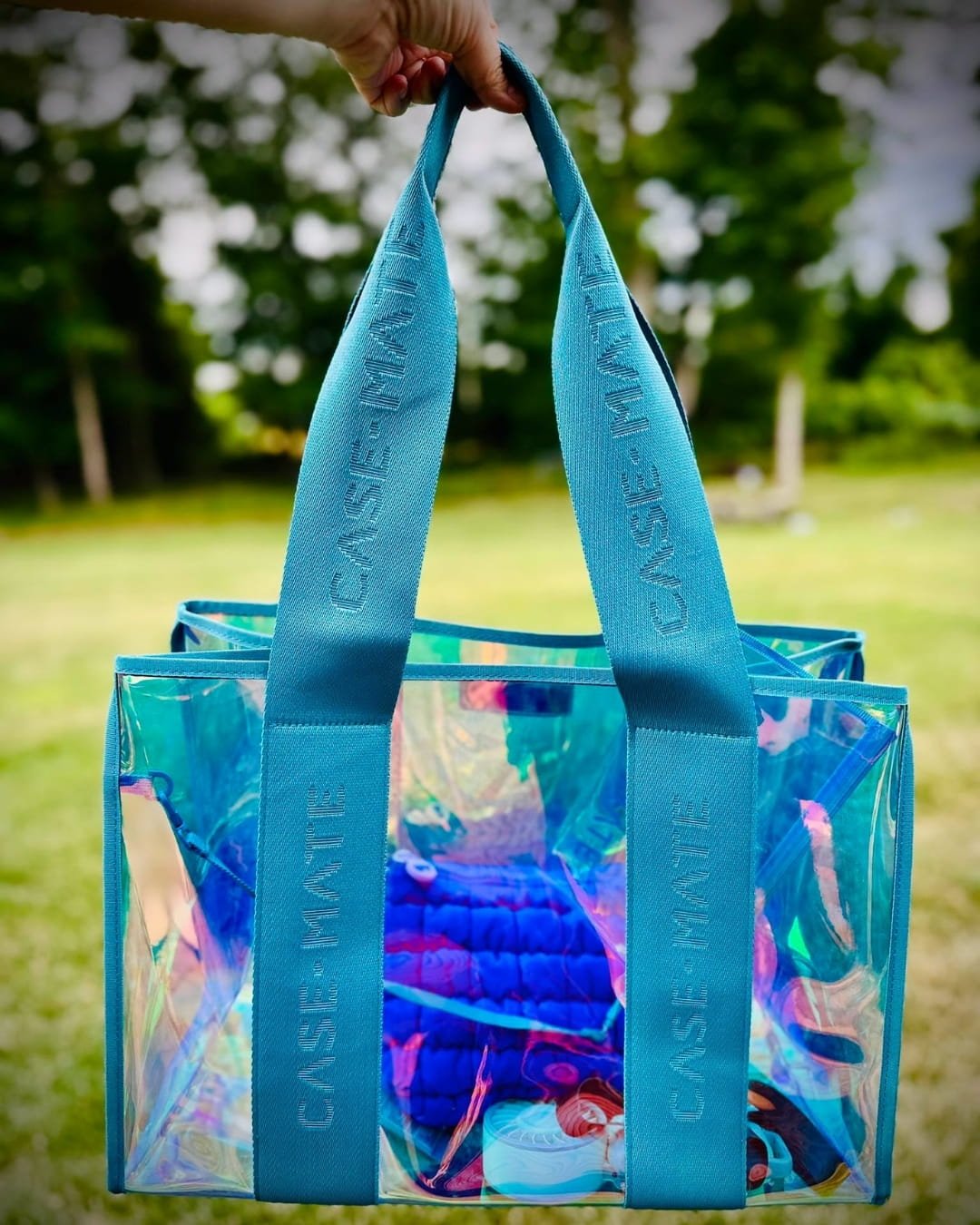 Iridescent clear Case-Mate jelly tote bag with teal branded straps held outdoors against a blurred green background