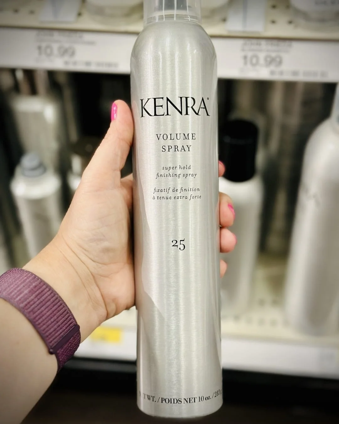 Hand holding a 10 oz. can of Kenra Volume Spray 25 with a metallic silver finish, displayed in a retail store aisle with shelves of hair products in the background.