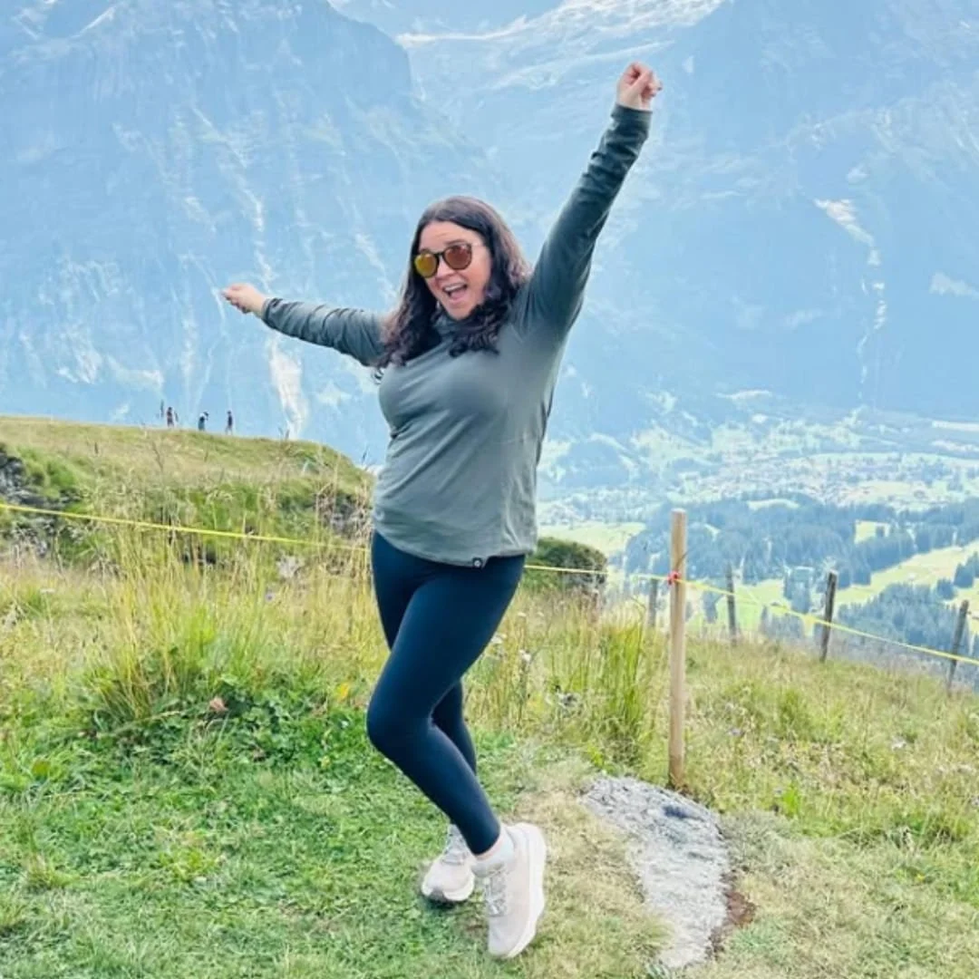 Nicole, an adventurous traveler, joyfully posing on a grassy hillside with arms raised, surrounded by a stunning mountain landscape and clear blue sky.