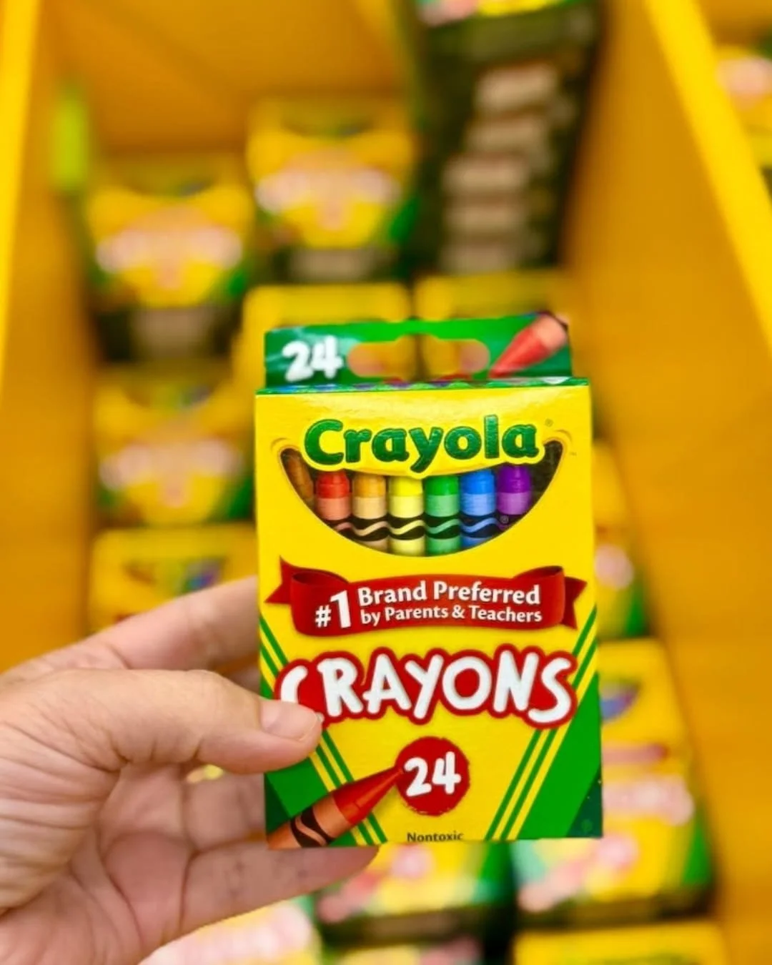 Hand holding a 24-count box of Crayola crayons with colorful crayon tips visible through the front window in a store aisle