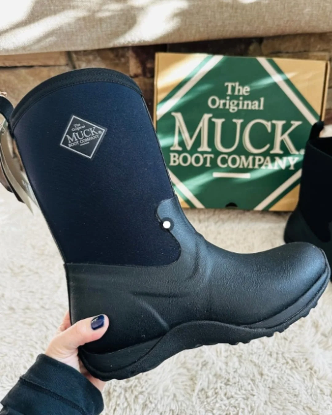 Navy blue and black waterproof Muck Women's Hunter Tall boot with a textured rubber sole, held by a hand with dark nail polish, displayed on a shaggy white rug with the original green and tan Muck Boot Company packaging in the background.