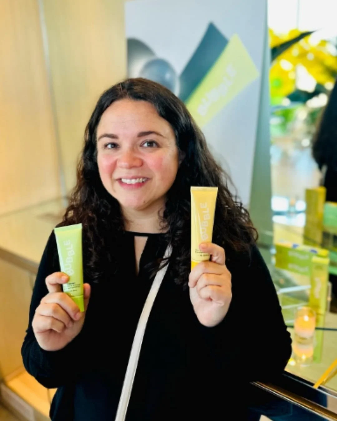 Nicole smiling and holding two BUBBLE skincare product tubes, one light green and one yellow, with a branded promotional background featuring the BUBBLE logo.