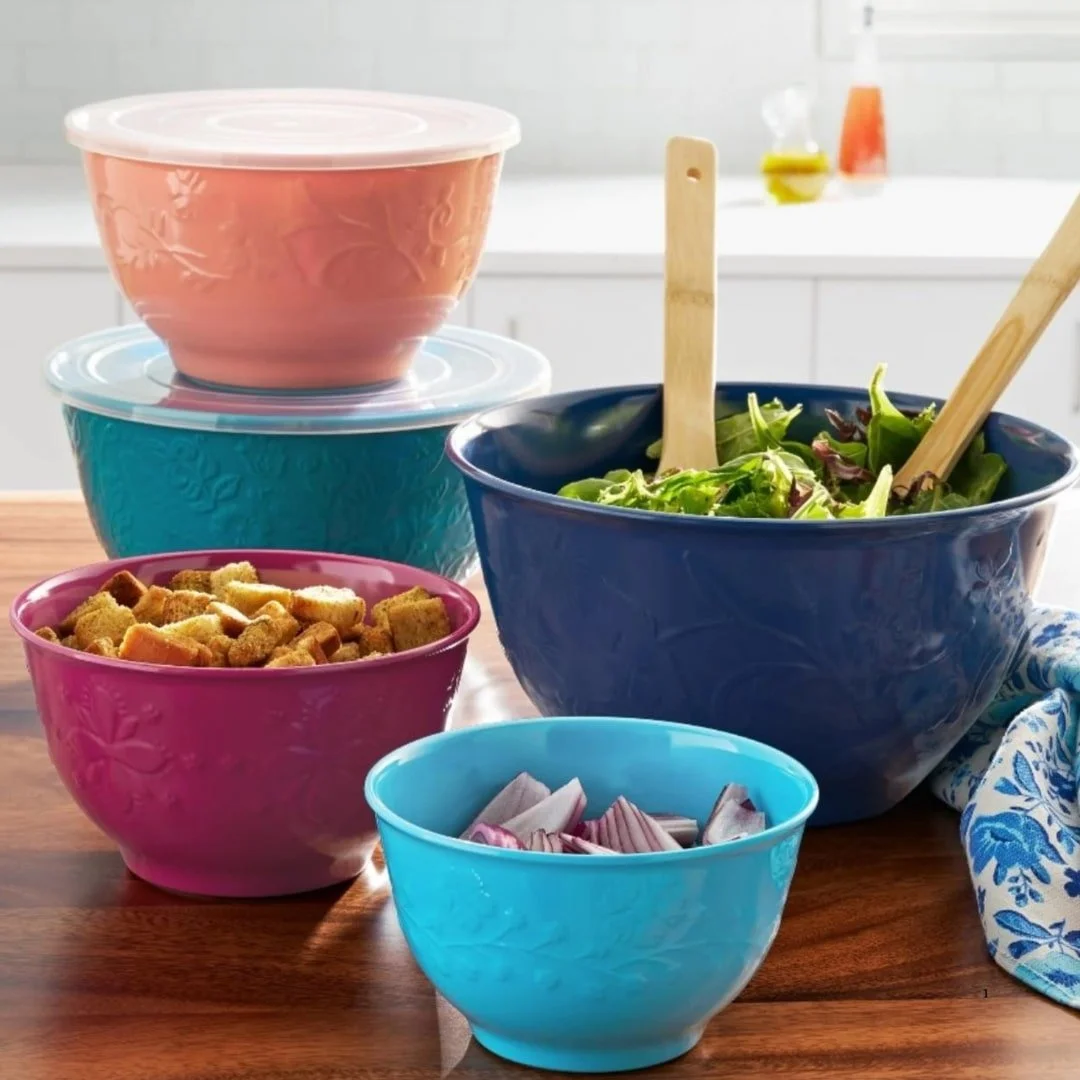 Set of five colorful embossed mixing bowls in peach, teal, magenta, dark blue, and cyan, displayed on a wooden table with food items like salad, croutons, and red onions.