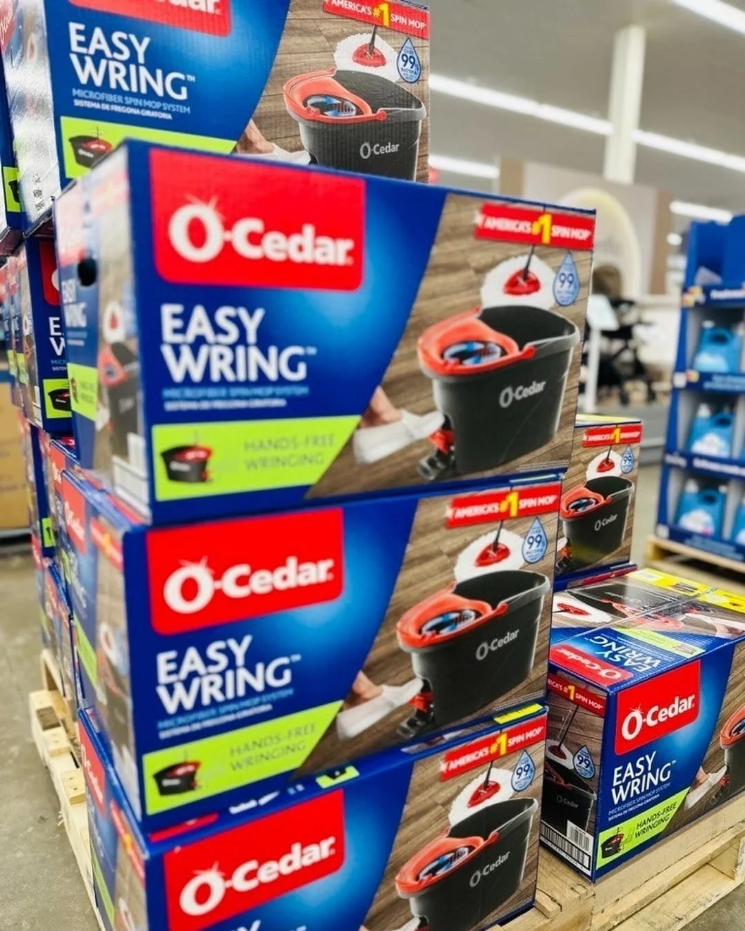 Stacked boxes of O-Cedar EasyWring Microfiber Spin Mop Systems on a wooden pallet in a retail store, highlighting features like hands-free wringing and 99% bacteria removal with water.
