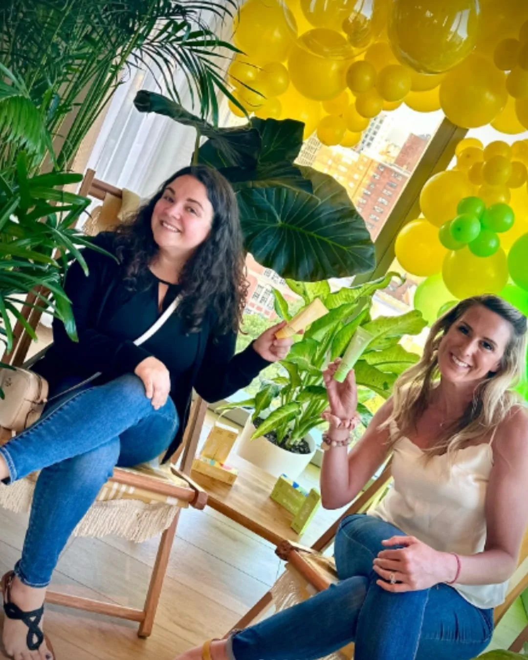 Nicole and Jenna seated in wooden chairs, smiling and holding yellow and green beauty product tubes, surrounded by lush plants and a yellow-green balloon arch at a product launch event.