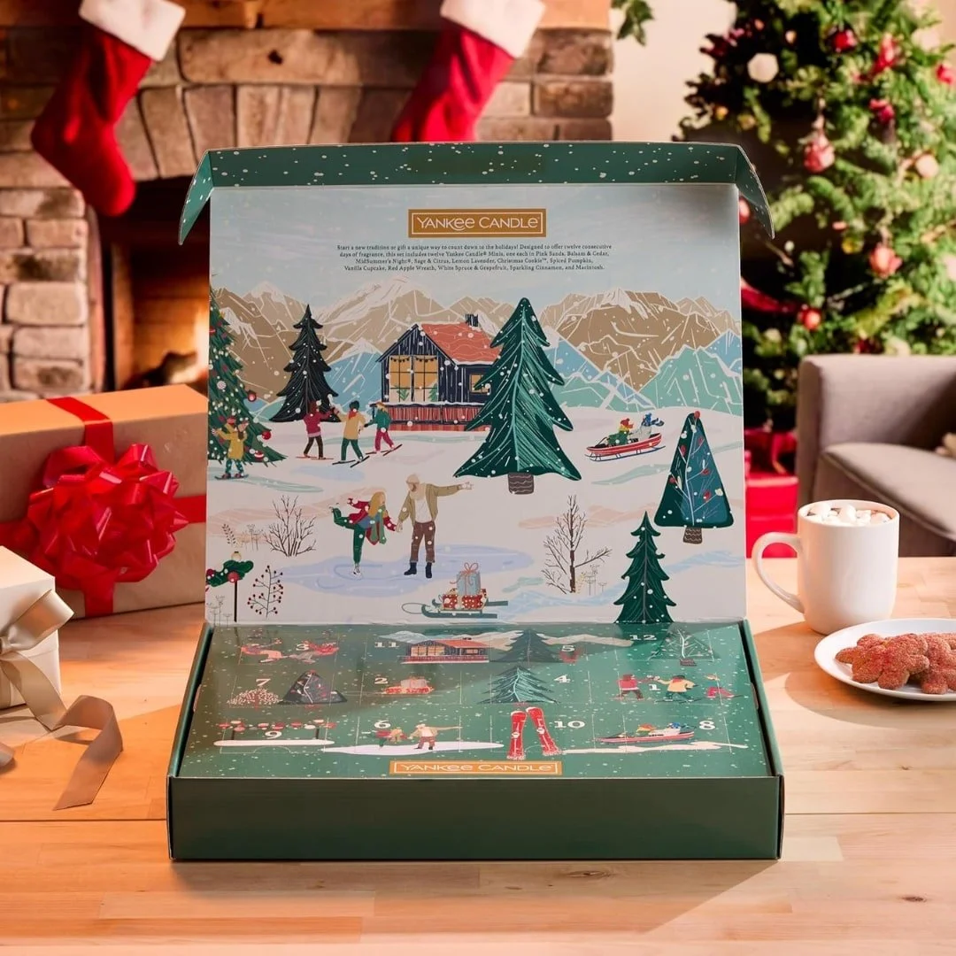 Festive Yankee Candle advent calendar featuring 12 mini candles in holiday-inspired scents, displayed on a wooden table with Christmas decorations, gifts, hot chocolate, cookies, and a cozy fireplace in the background.