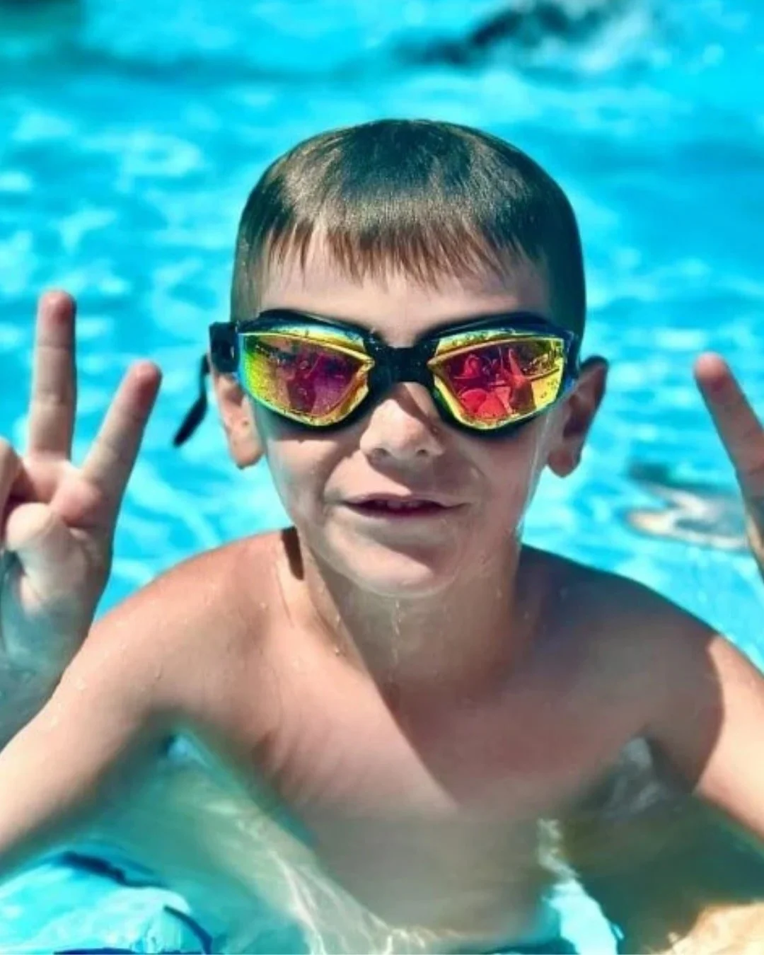 Smiling boy in pool wearing goggles, making peace signs.