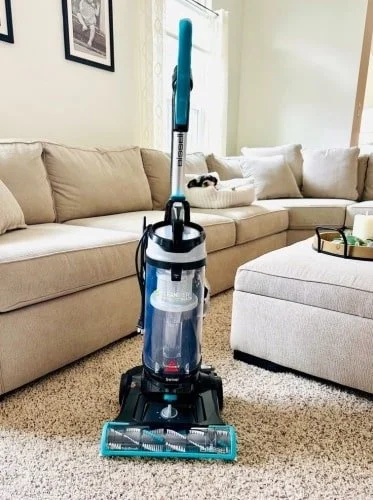 Bissell vacuum cleaner with teal and black design on a light shaggy carpet in a bright living room, featuring a beige sectional sofa, ottoman with a tray, and framed wall decor.