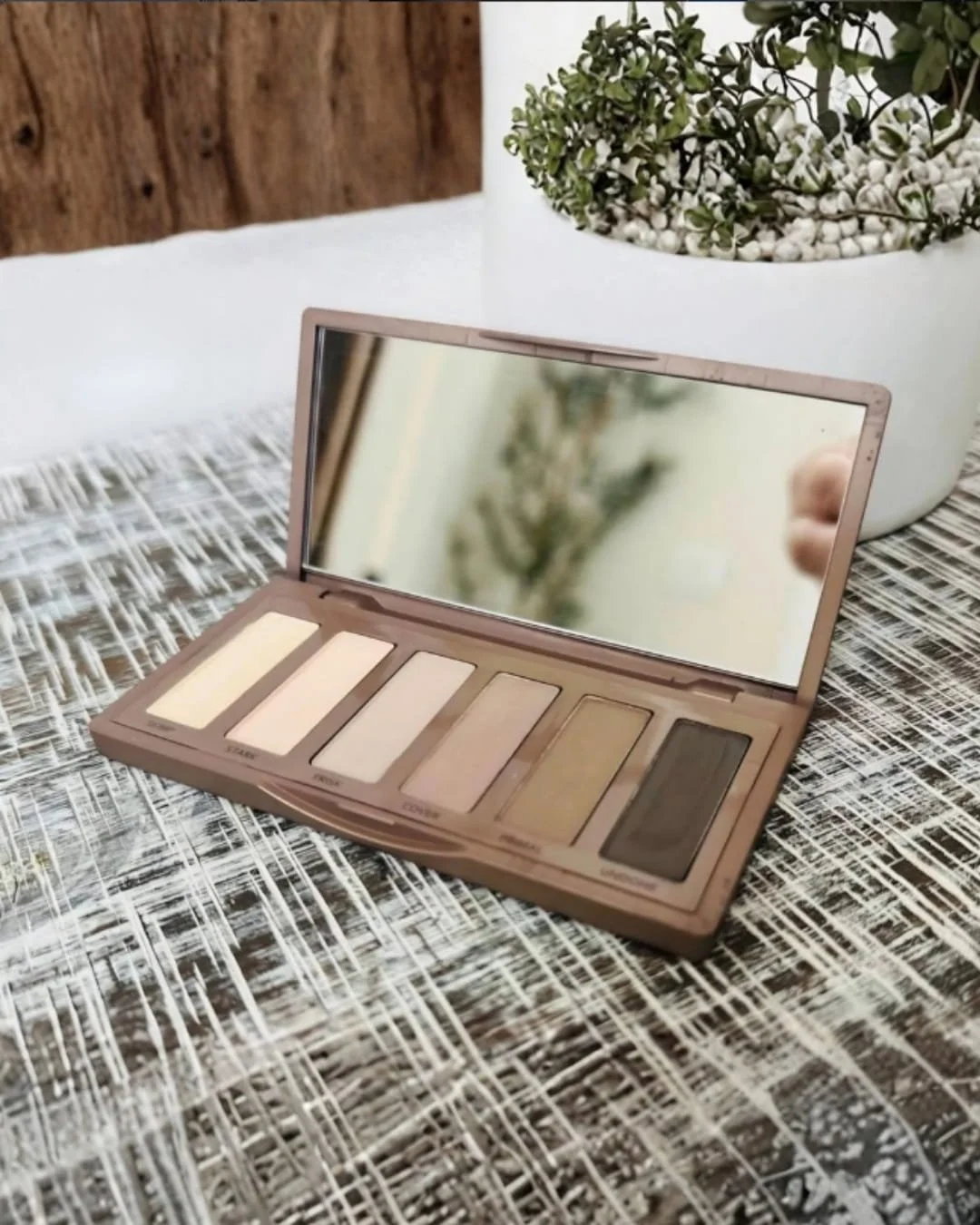Open mini eyeshadow palette with six neutral matte shades and a built-in mirror, displayed on a textured surface beside a small potted plant