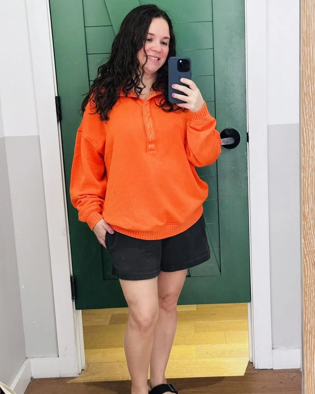 Woman taking a mirror selfie wearing a bright orange oversized sweatshirt and black Aerie Vacay shorts, standing in front of a green barn-style door with light wood flooring and neutral walls.