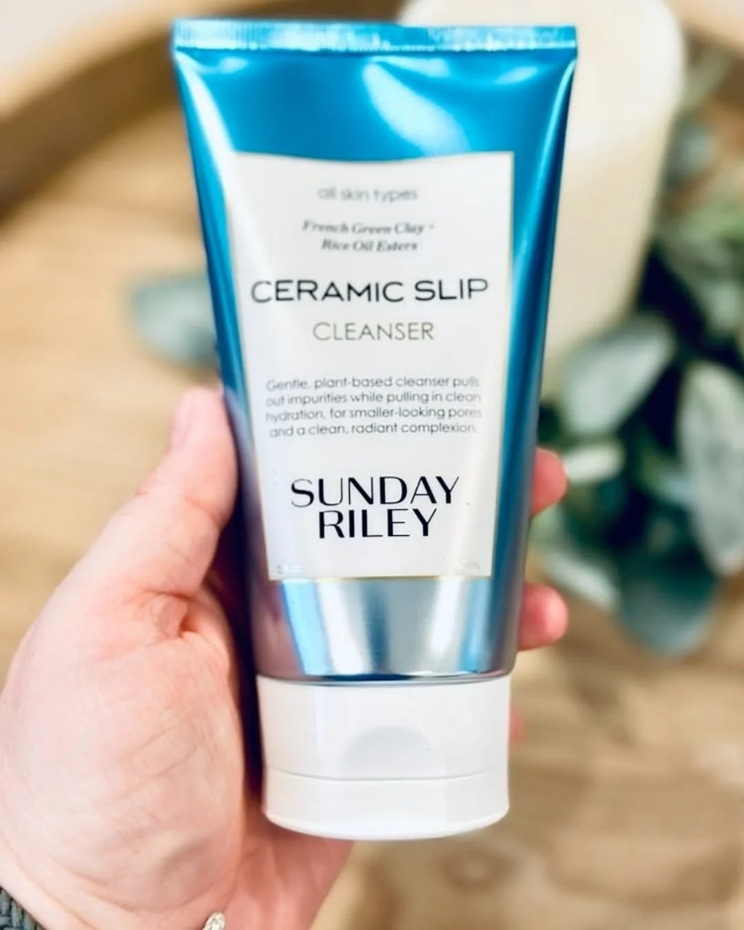 Sunday Riley Ceramic Slip Cleanser tube held in hand with candle and greenery in background
