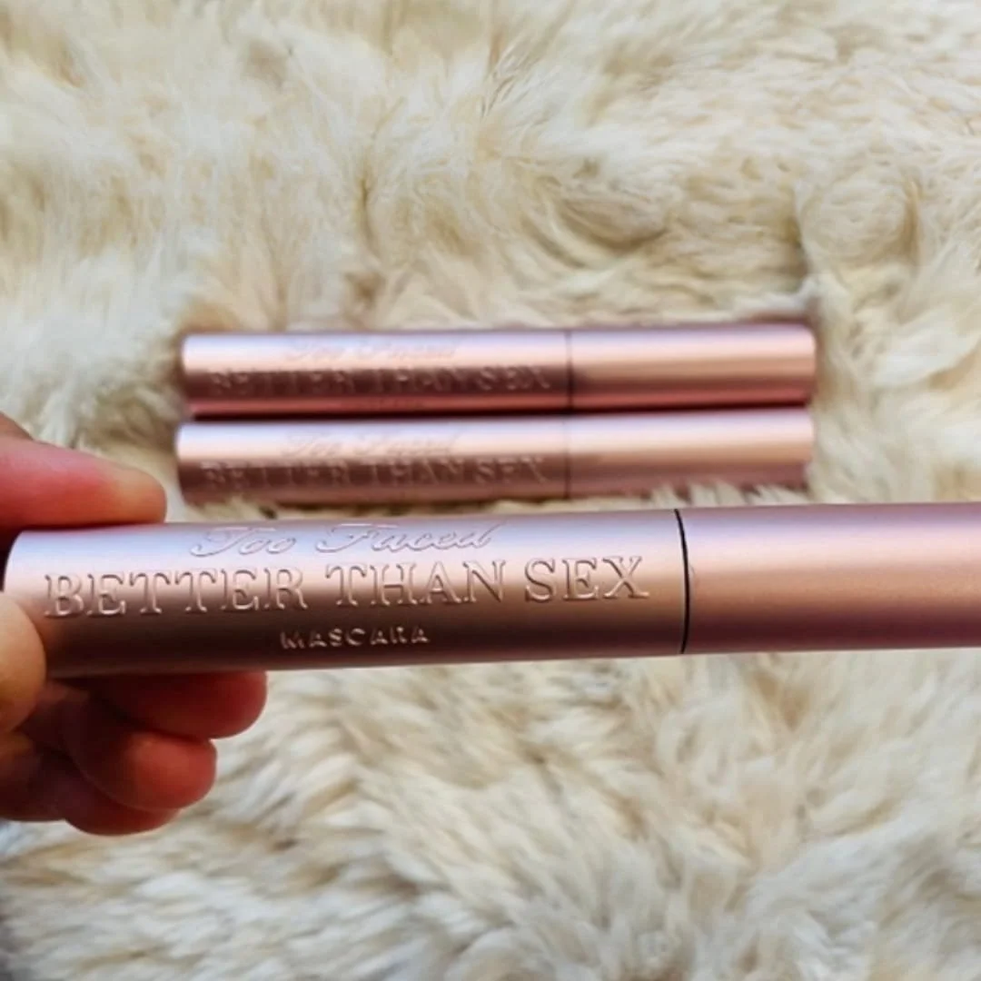 Three tubes of Too Faced Better Than Sex Mascara with metallic rose gold packaging, one held in a hand and two lying on a soft white furry background, showcasing the embossed branding.