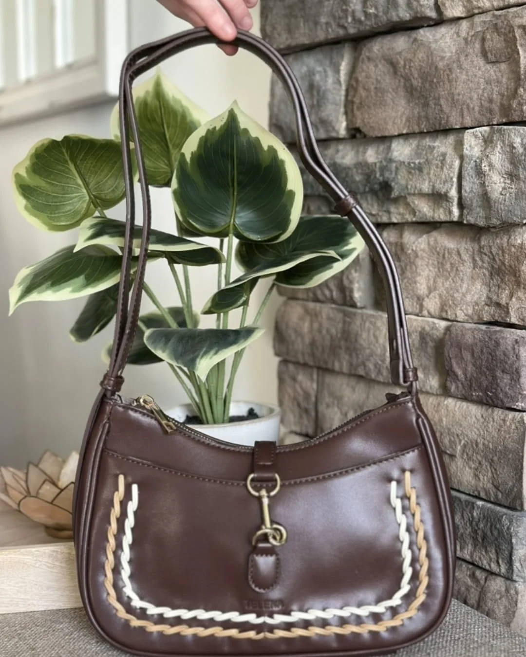 Dark brown Telena shoulder bag with cream and tan braided detailing, gold-tone hardware, and adjustable strap, displayed against a backdrop of a green potted plant and a rustic stone wall.