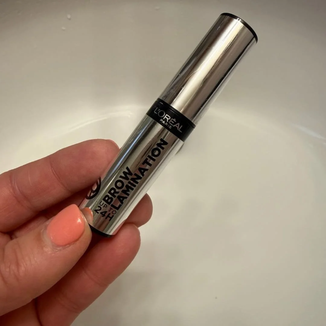 L'Oréal Paris Brow Lamination gel in a sleek metallic silver tube with '24H' long-wear claim, held by a hand with coral nail polish against a light gray background.