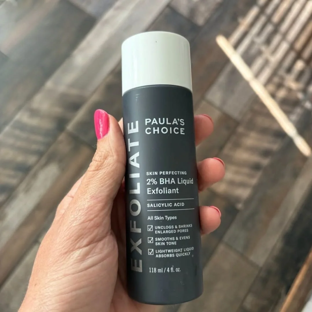 Hand holding Paula’s Choice Skin Perfecting 2% BHA Liquid Exfoliant bottle with white cap against a wooden background