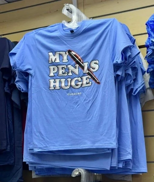 Funny light blue t-shirt with MY PEN IS HUGE slogan and pen graphic, souvenir from Curaçao, displayed on a hanger in a retail store with folded shirts in the background.