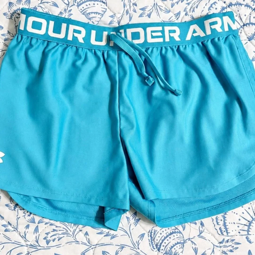 Bright turquoise Under Armour girls' athletic shorts with elastic logo waistband and drawstring, laid flat on a patterned quilt