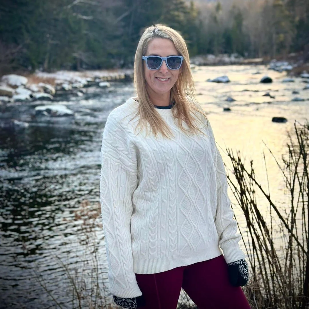 Jenna, content creator for Nicole Fabulous Finds, standing by a winter river in a cream cable-knit sweater, maroon leggings, and sunglasses, with a snowy forest and golden light in the background.