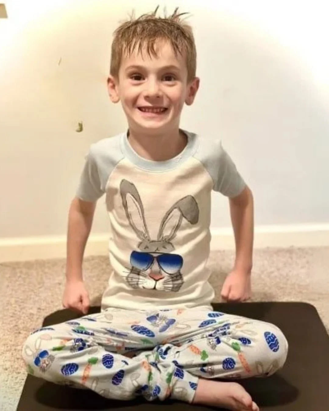 Smiling young boy sitting cross-legged on a dark mat, wearing Easter-themed pajamas with a bunny graphic and colorful egg and carrot patterns, against a light-colored wall.