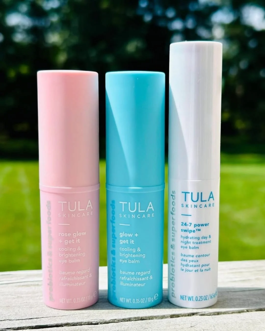 Three TULA Skincare eye balm sticks—pink Rose Glow + Get It, blue Glow + Get It, and white 24-7 Power Swipe—arranged on a wooden surface with a blurred green outdoor background, highlighting their vibrant and clean design.