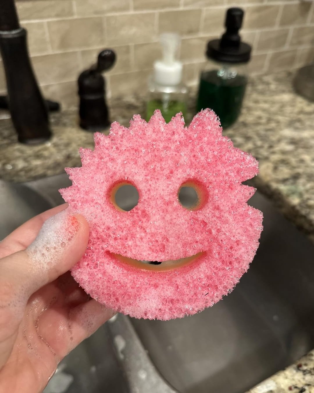 Pink Scrub Mommy sponge with a smiling face design, held by a soapy hand over a modern kitchen sink with soap dispensers and a granite countertop in the background.
