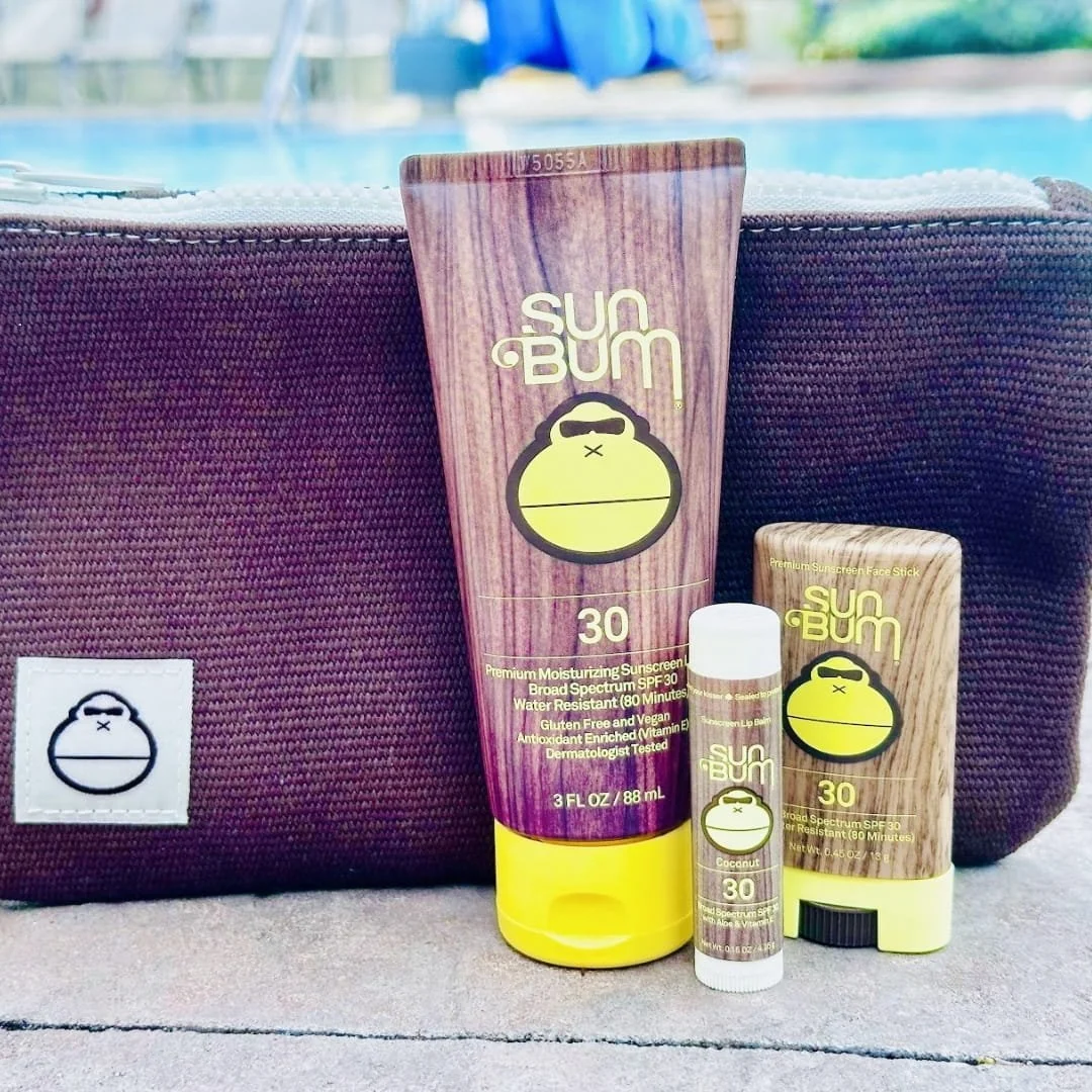 Sun Bum SPF 30 travel set featuring sunscreen lotion, lip balm, and face stick with a brown toiletry bag, displayed on a stone surface by a poolside with a sunny outdoor backdrop.