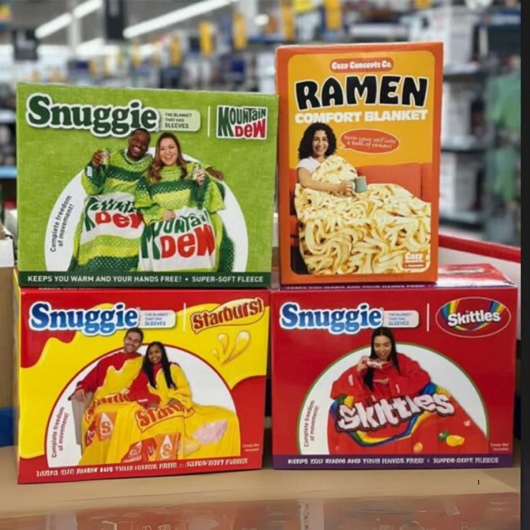 Product boxes for Snuggie and Ramen comfort blankets, showcasing food and drink-themed designs like Mountain Dew, Starburst, and Skittles, on a retail shelf.