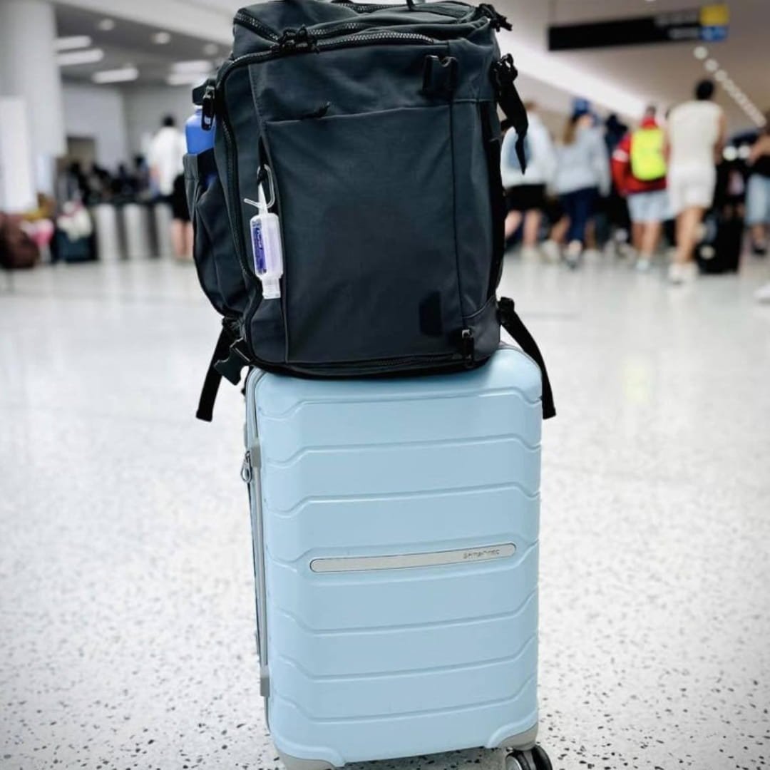 Samsonite light blue hardside carry-on luggage with a black backpack and hand sanitizer attached, placed in an airport setting with blurred travelers in the background.
