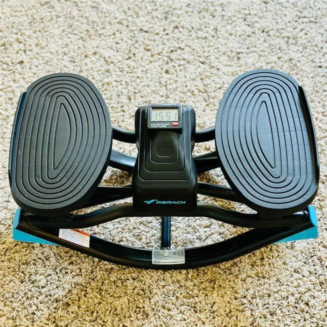 MERACH mini-stepper exercise machine with black textured foot pedals, a central LCD display showing workout metrics, and bright blue end caps, placed on a beige shag carpet for home fitness use.