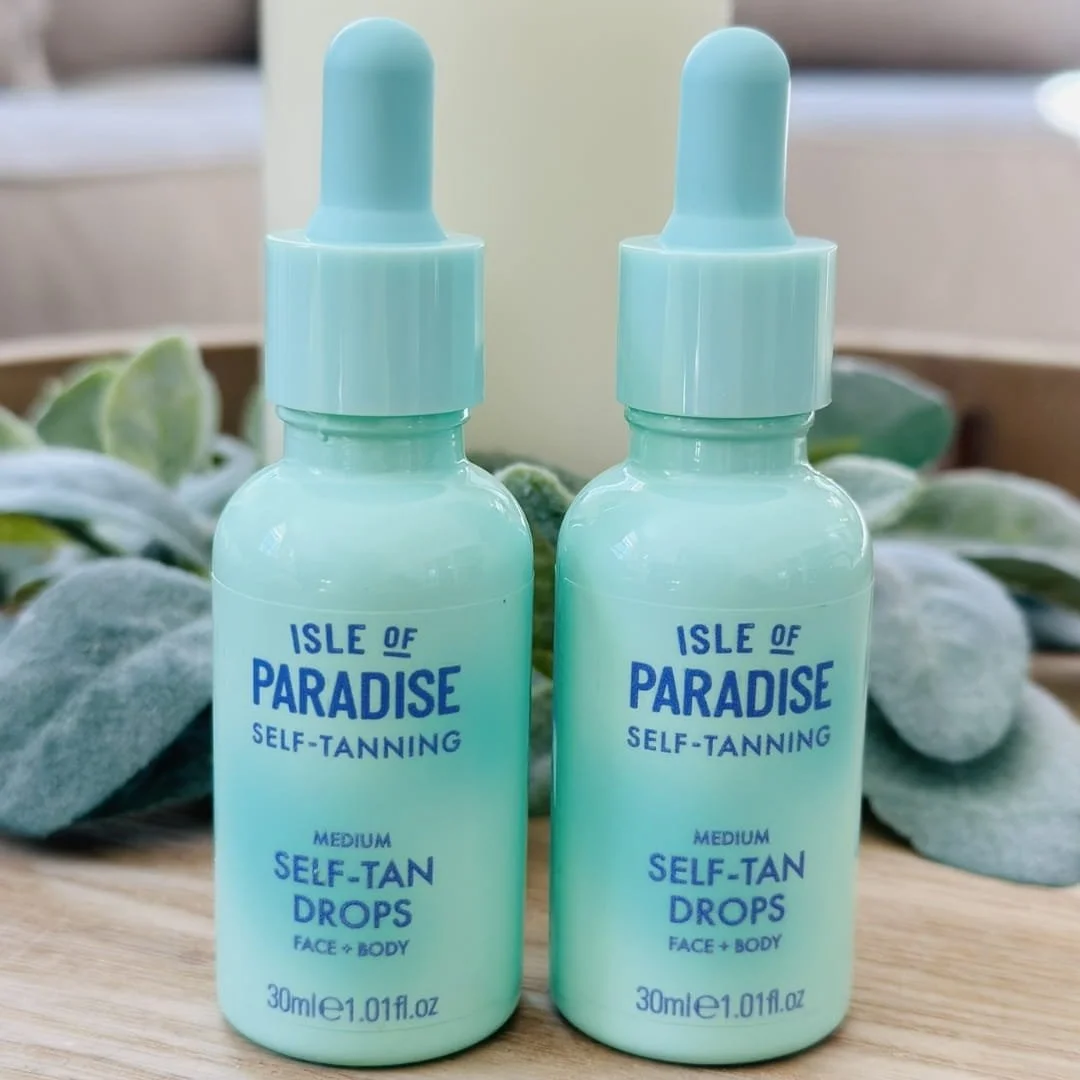 Two mint green bottles of Isle of Paradise Medium Self-Tan Drops for face and body, displayed on a wooden surface with soft greenery in the background.